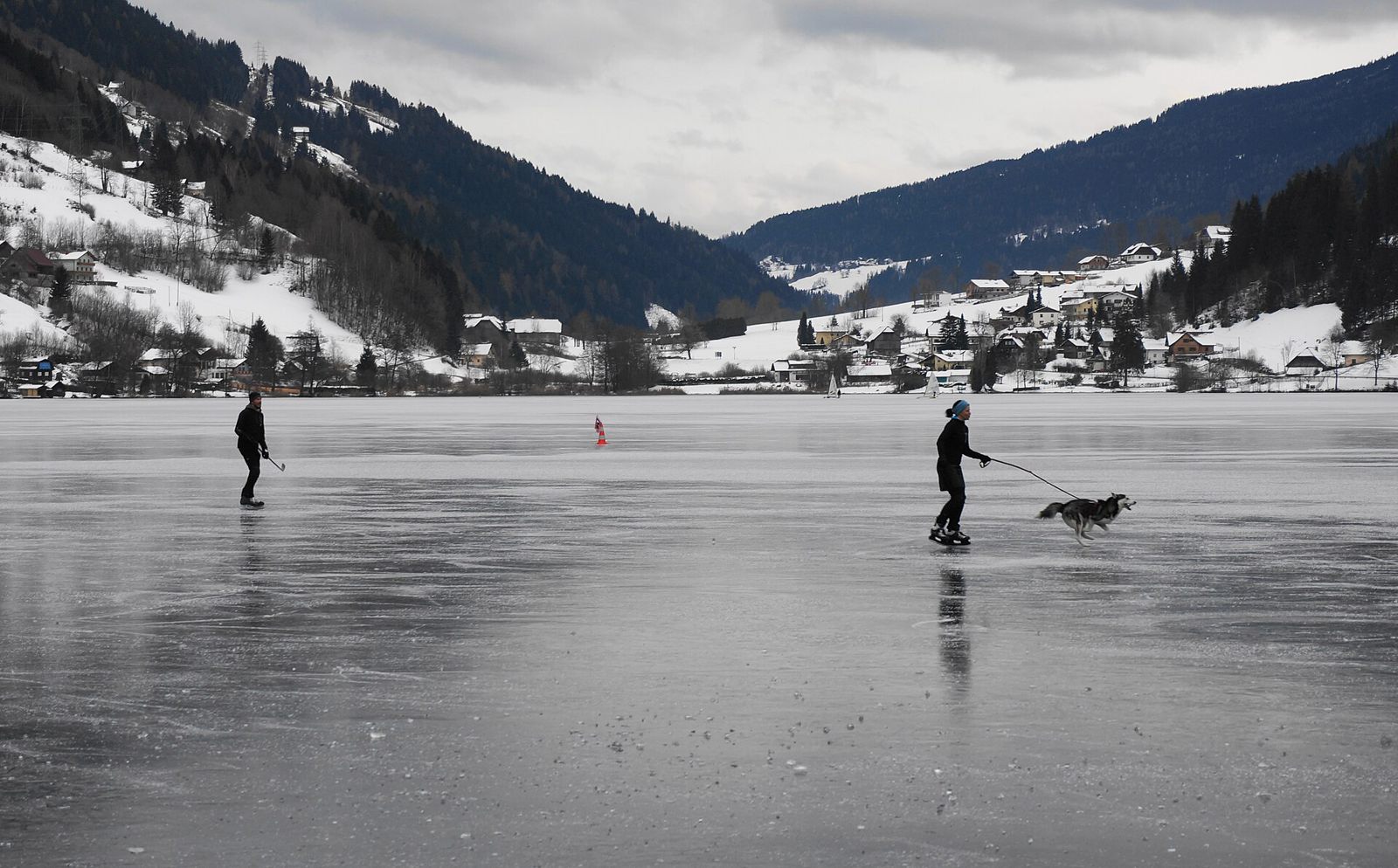 Ice skaters and a dog crossing frozen Brennsee in front of a snowy village